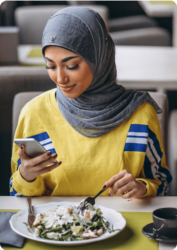 A muslim woman in hijab eating food at a restaurant while looking at her phone and smiling
